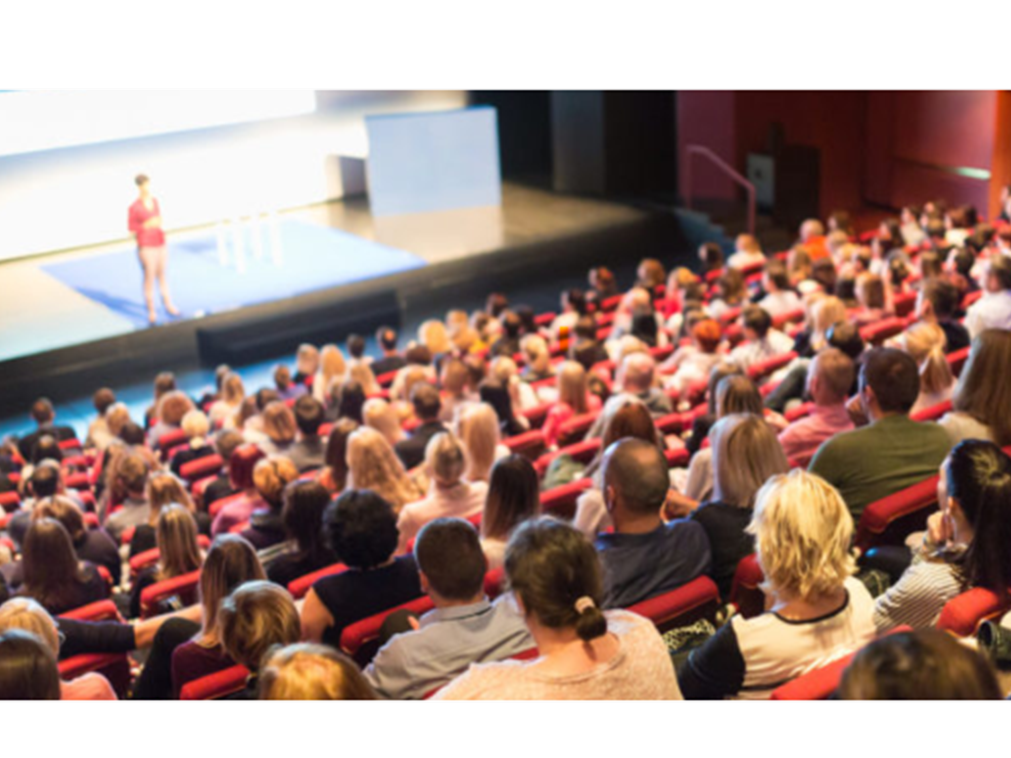An image from the back of a lecture hall shows a large audience watching a speaker on stage. 