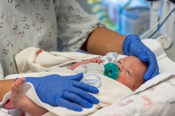 This image shows a healthcare professional gently cradling a newborn baby in a hospital bed who is wrapped in a blanket with "back is best" embroidered on it, promoting safe sleep practices. 