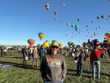 Dr. Elena Griego stands outside, with a group of hot air balloons rising from a field behind her. 